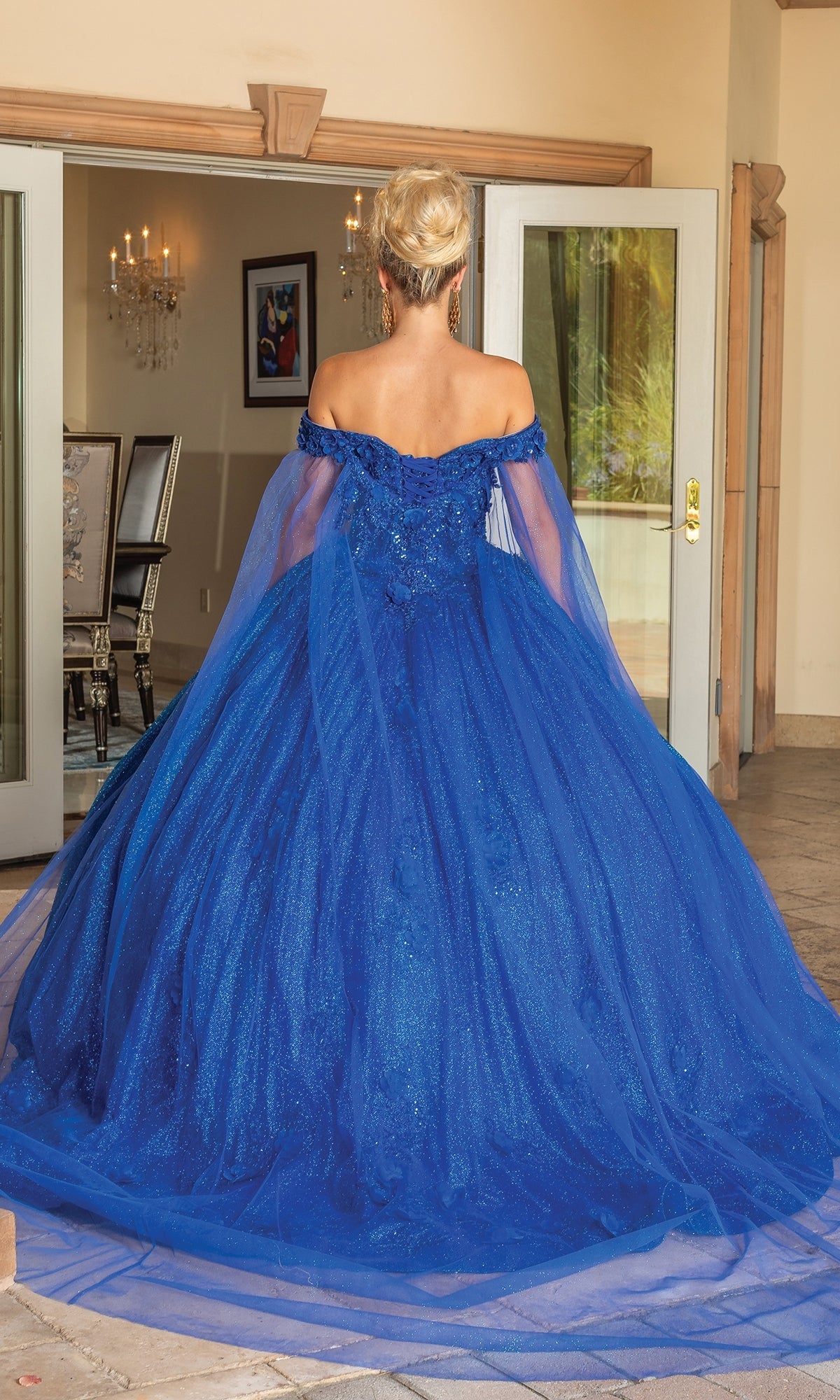 A person with blonde hair in an updo wears the Quinceanera Dress 1718 By Dancing Queen, a royal blue off-the-shoulder gown with a sweetheart neckline, standing indoors with their back to the camera.